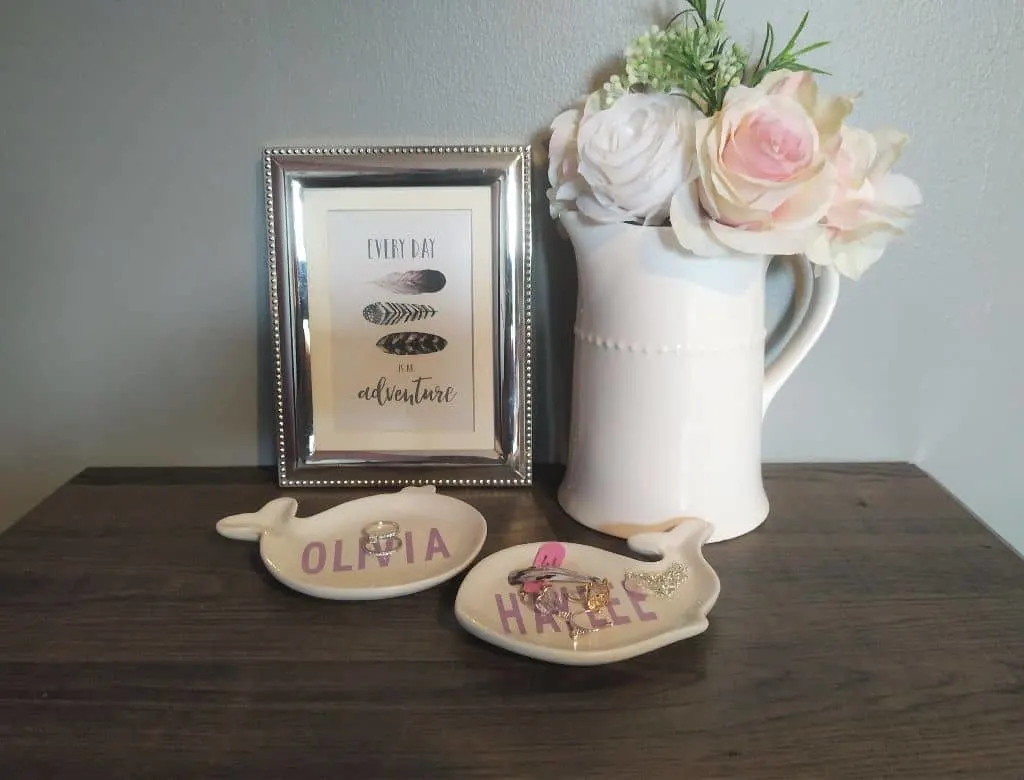 Whale shaped trinket dishes next to a picture frame and a pitcher full of flowers on a wooden desk.