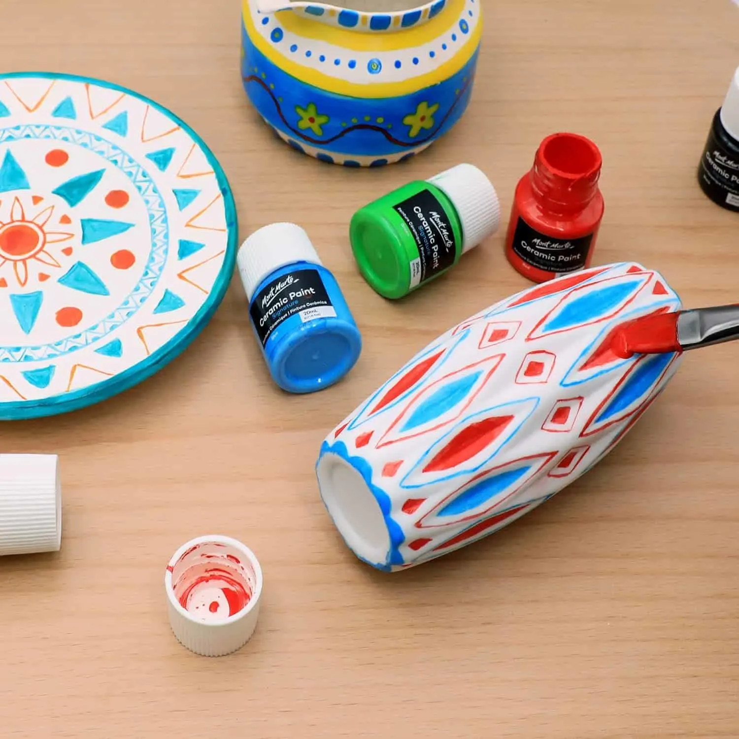 ceramic paint jars on a table with a plate painted with a triangle motif, a vase laying on its side and being painted with paint brush and a vase with floral and dot details painted on it.