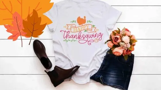 a pair of boots, a t-shirt with a turkey sublimation design and the words happy thanksgiving spread on a wooden floor next to a folder pair of jean shorts and a bouquet of roses