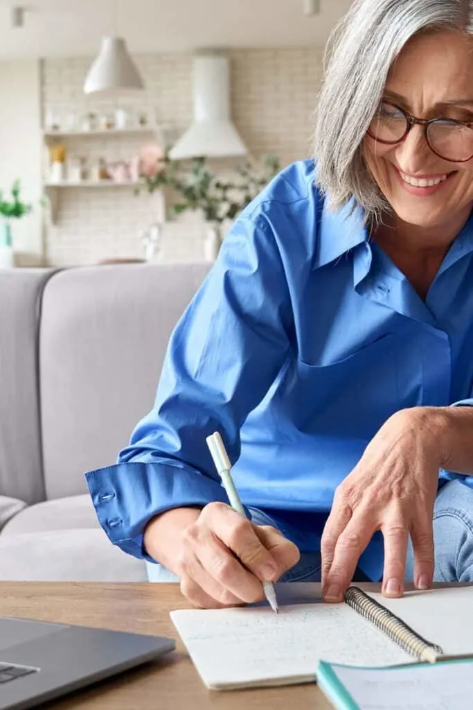An elderly woman writing in a notebook and her laptop is open on a table in front of her.