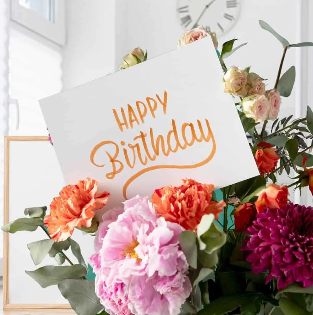 A birthday card in the middle of a bouquet of carnations. There is a window, a picture frame and a clock on the wall in the background.