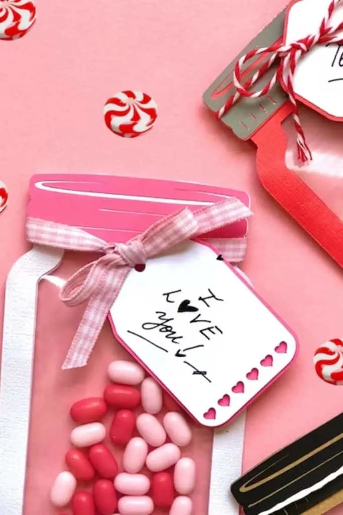 The image features a close-up of a pink mason jar lying on its side, filled with a variety of pink and red candies, on a matching pink background. The jar is adorned with a gingham ribbon tied into a neat bow. Attached to the jar is a white gift tag with the handwritten phrase "LOVE you!" accompanied by small black hearts. Scattered around the jar are peppermint candies, adding to the festive Valentine's Day theme.
