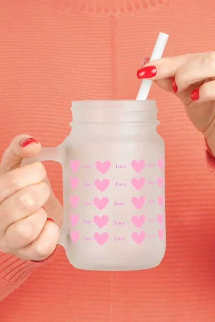 A frosted mason jar mug with a handle, decorated with rows of pink hearts and the word "love" in a repeating pattern. A person's hand is holding the mug, showcasing red nail polish that matches the hearts' color, while a white straw is inserted through the mug's lid, suggesting a ready-to-enjoy beverage. The background is a textured coral hue, complementing the mug's romantic motif.