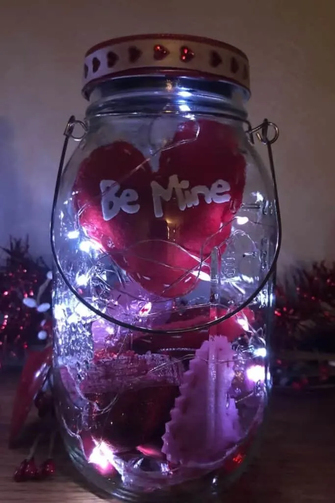 The image features a clear mason jar with a wire handle, illuminated from within by a string of fairy lights, creating a warm glow. Inside the jar, a red heart with the words "Be Mine" stands out against the light. The jar's lid is adorned with a Valentine-themed design featuring small red hearts. The romantic ambiance is enhanced by the soft lighting and the festive, love-inspired decorations inside the jar, making it a charming piece for Valentine's Day.