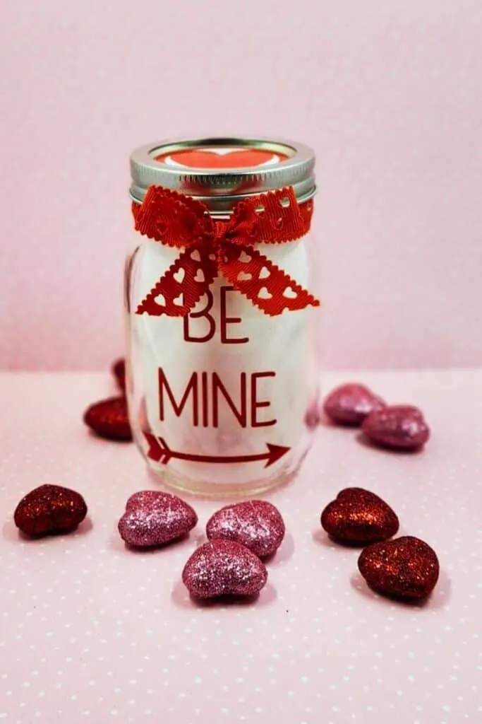 The image shows a clear mason jar with a red ribbon tied in a bow around the neck and adorned with lace detailing. The jar has the words "BE MINE" in brown letters, giving it a charming Valentine's Day feel. Scattered around the jar are glitter-covered, heart-shaped chocolates in shades of red and purple, all resting on a soft pink background with a subtle polka-dot pattern. The overall look is sweet and romantic, perfect for a Valentine's Day gift.