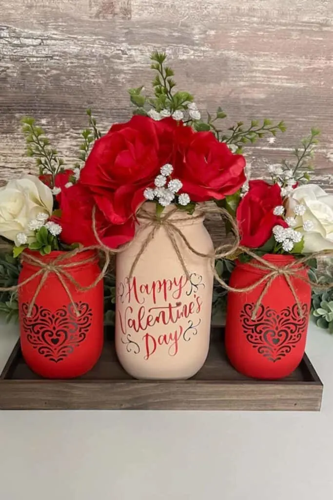 The image shows a trio of painted mason jar vases on a wooden tray against a rustic wooden backdrop. The central jar is cream-colored with "Happy Valentine's Day" scripted in red, flanked by two red jars featuring elegant black scrollwork designs. Each jar is wrapped with a natural twine bow and is bursting with vibrant red and delicate white roses, creating a festive and romantic Valentine's Day display.