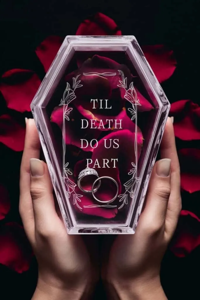 a unique and gothic-inspired wedding ring box. The box is shaped like a small, transparent coffin, held gently in both hands against a dark background. Inside the coffin, two silver wedding rings rest on a bed of deep red rose petals. The front of the coffin is adorned with delicate white line art and floral details framing the text "TIL DEATH DO US PART" in a bold, vintage-style font. The overall atmosphere of the image is romantic with a dark, elegant twist, perfect for an unconventional wedding theme.