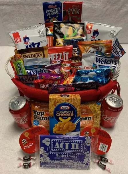 A whitle wicker basket lined with a red cloth and filled with popcorn, fruit snacks, potato chips, mini cereal boxes, mac and cheese, coco cola and ramen noodles