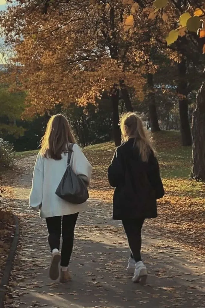 Two women walking along a tree-lined path in a park during autumn. The ground is scattered with fallen leaves, and the trees above are ablaze with golden and orange foliage. Both women are dressed in cozy, oversized jackets and leggings, carrying bags over their shoulders, enjoying the peaceful, crisp fall weather. The soft sunlight filters through the trees, casting a warm glow over the scene, evoking a sense of calm and friendship during a seasonal stroll.