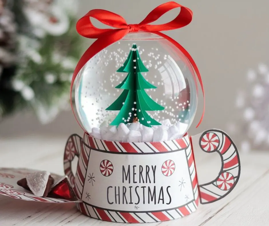 A DIY Christmas gift featuring a decorative mini snow globe with a hidden treat compartment. The clear plastic globe contains a 3D green Christmas tree surrounded by white foam chunks resembling snow and is topped with a red ribbon bow. The base displays a "Merry Christmas" message with red-and-white Hershey kisses candy.