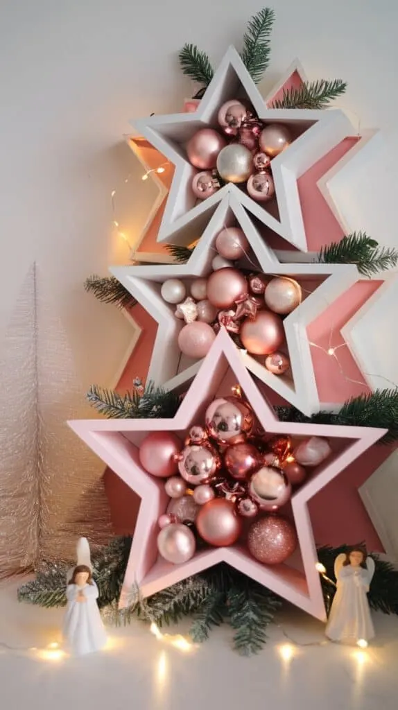 This image features a decorative, festive display arranged on a flat surface, like a shelf or table. It includes a tiered set of three-dimensional, nested star-shaped shelves, painted white and arranged in a staggered, stacked fashion, forming a star-like structure. The stars are filled with pink and metallic-colored ornaments, including Christmas baubles and small decorative stars. Some soft fairy lights are woven around the stars, adding a warm, illuminated glow.