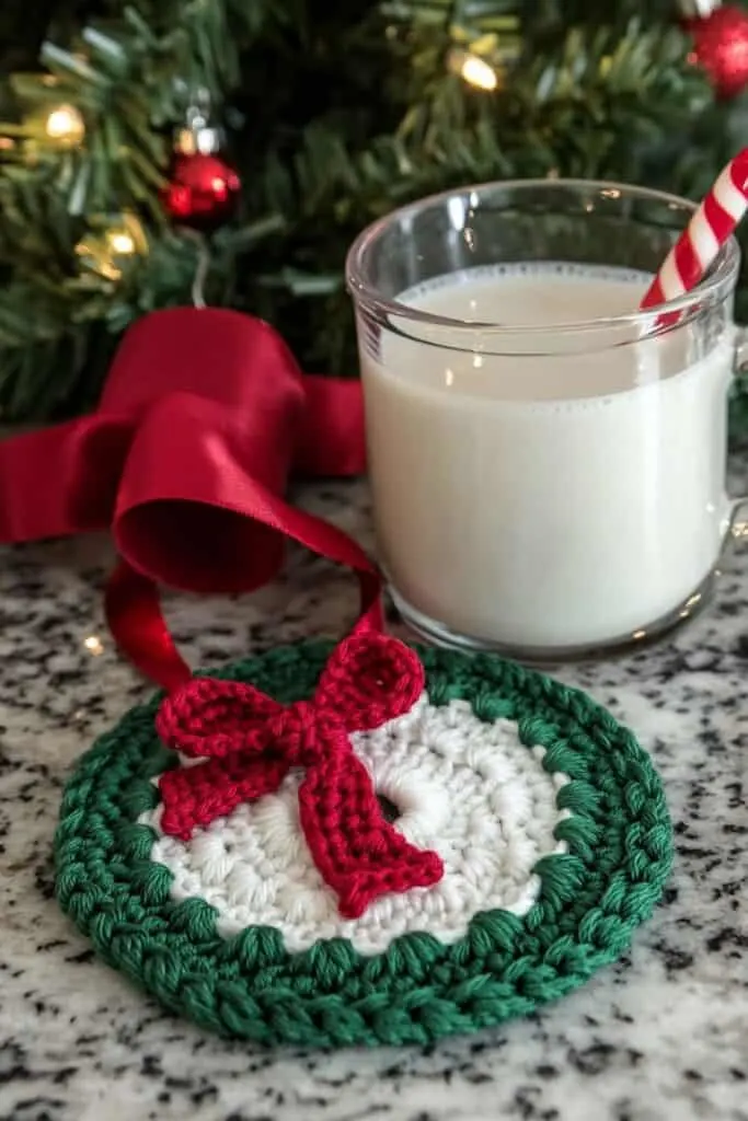 A crocheted coaster designed in a festive holiday theme. The coasters are circular with a white center and a green scalloped border. A red ribbon detail is woven into the design, tying into a small bow. A small glass mug filled with eggnog is placed next to the coaster. There is also a Christmas tree in the background, enhancing the festive look. The setup is on a marble countertop in a kitchen, creating a cozy holiday atmosphere.