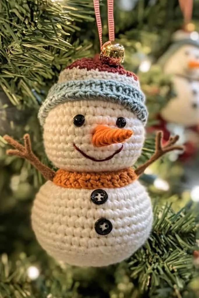 A photo of a crocheted snowman used as a Christmas ornament. The snowman is hanging on a Christmas tree branch. The snowman has a smiling face, a carrot nose. The snowman's body has buttons and pipe cleaner twigs for its arms. The tree has 
 lights.