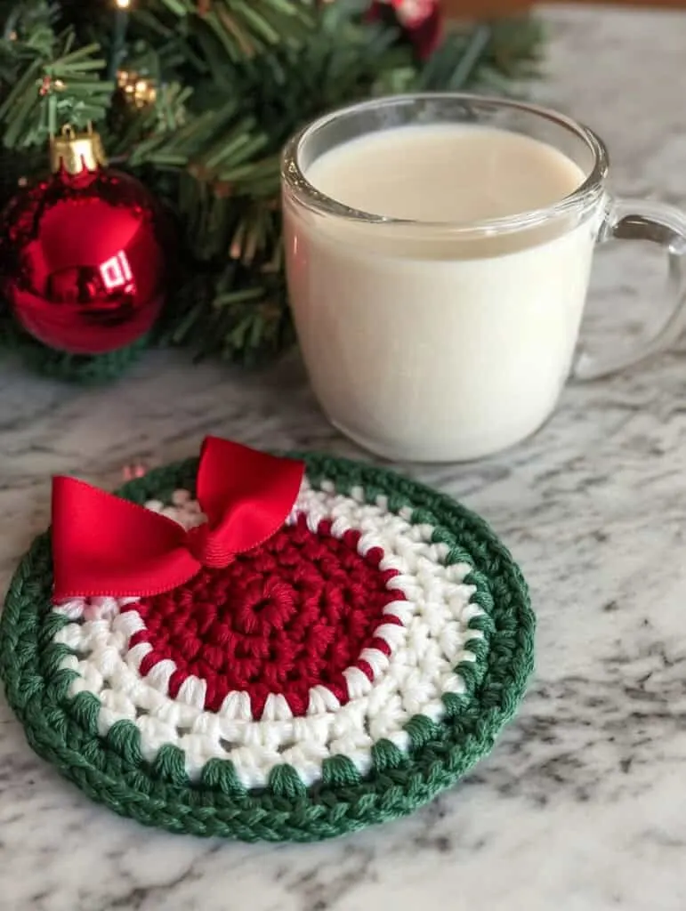 A crochet coaster designed in a festive holiday theme. The coasters are circular with a white center and a green scalloped border. A red ribbon detail is woven into the design, tying into a small bow. A small glass mug filled with eggnog is placed on 
 the coaster. There is also a Christmas tree in the background, enhancing the festive look. The setup is on a marble countertop in a kitchen, creating a cozy holiday atmosphere.