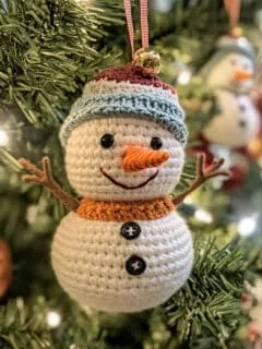 A photo of a crocheted snowman used as a Christmas ornament. The snowman is hanging on a Christmas tree branch. The snowman has a smiling face, a carrot nose. The snowman's body has buttons and pipe cleaner twigs for its arms. The tree has lights.